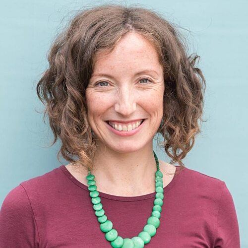 White woman with wavy brown bobbed hair, smiling wearing a green necklace over a burgundy top