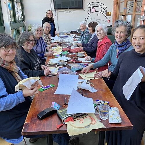 Group of women sat around a large rectangular table doing embroidery using wooden hoop frames. The table is covered in materials