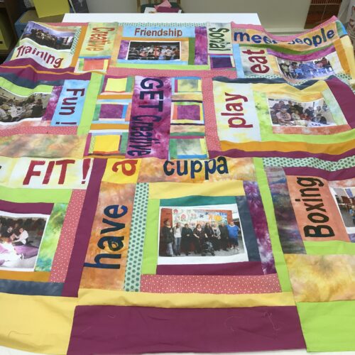 Large fabric quilt covering a table made up of squares in different colours. Some squares include words and others include photographs