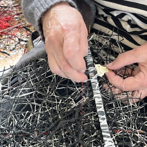 Hands wrapping a white thread around a thin pole that forms part of a larger black and white textile