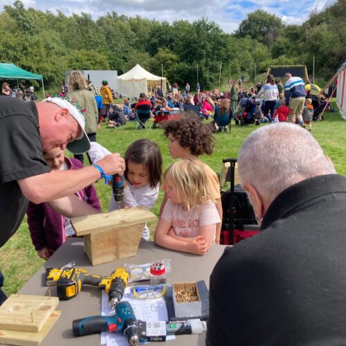 Two white men dressed in black working at a table outdoors showing young girls how to use tools