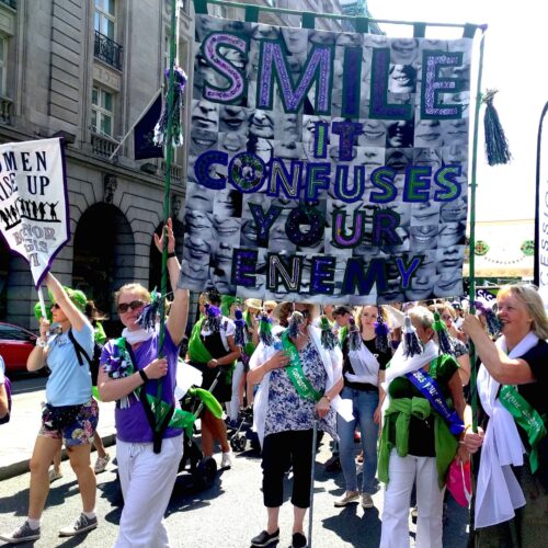 Group of women dressed in purple, white and green holding a large banner saying, smile it confuses your enemy