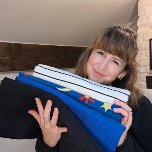 A young white woman with brown hair and smiling is holding a stack of different patterned materials in stripey blue and white, blue, and black