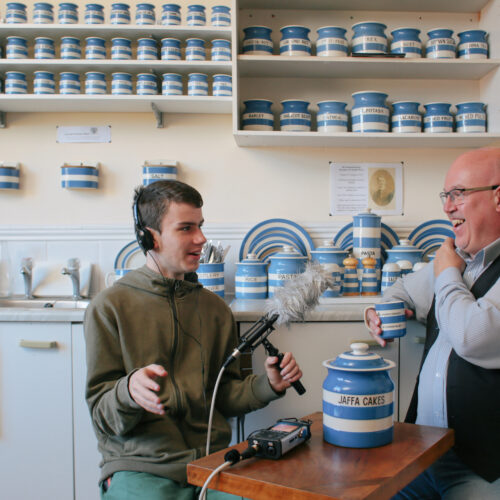 A young man wearing headphone is holding a microphone in front of an older bald man who is holding a blue and white stripey mug and laughing. They are sat in a kitchen surrounded by Cornish ware blue and white stripey crockery