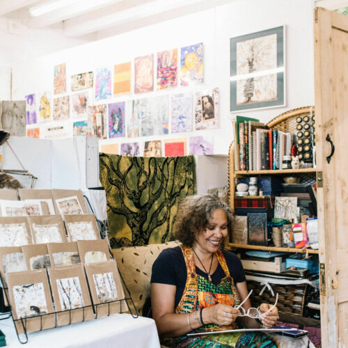 Woman in a brightly coloured dress sat in a booth surrounded by a rack of cards, paintings on the wall and a shelf unit full of bits and bobs