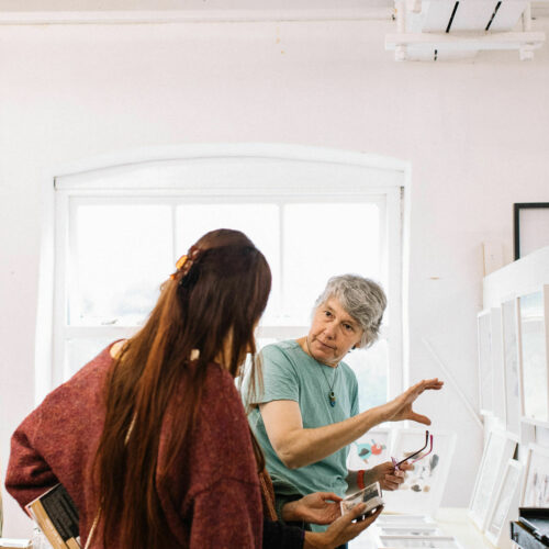 White woman wearing a green t-shirt explains her artwork to a woman with her back to the camera with long hair, in front of a table