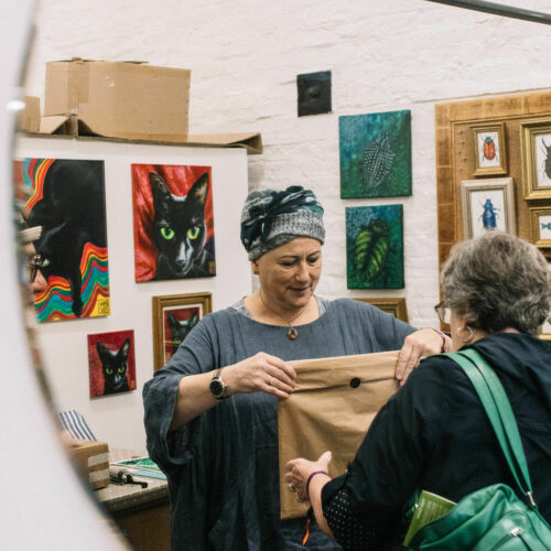 White, female artist wearing a turban standing in front of walls of paintings, handing over a wrapped artwork to a woman carrying a green bag over her shoulder