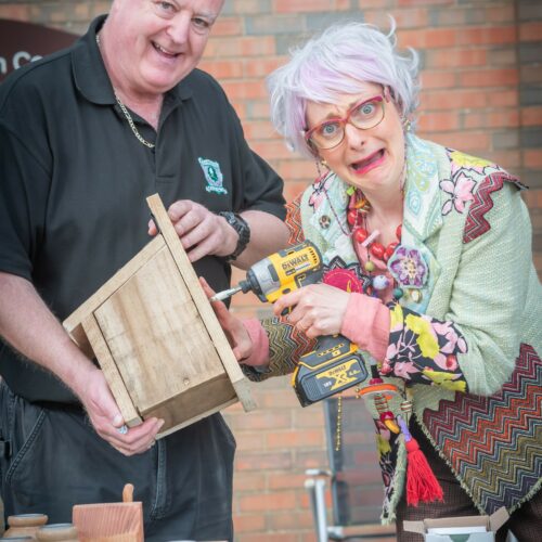 White man dressed in black holding a wooden box as a white woman with brightly coloured clothing, glasses and hair holds a drill against the box, with a scared look on her face