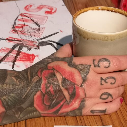 A tattoed hand with the date 1963 across the fingers, and a rose running from the hand onto the arm, is holding a brown ceramic mug. Next to the jug is a hand-drawn picture of a large spider in black on paper