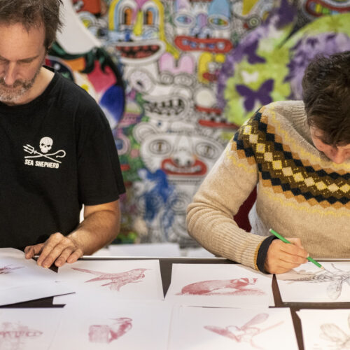 Two people sat drawing creatures on white paper. A man with a grey beard, wearing a black t-shirt is drawing with a red pen, and the other person, wearing a traditional Shetland style woollen jumper with a pattern around the yoke, is drawing with a black pen. Lots of red drawings are visible on the table