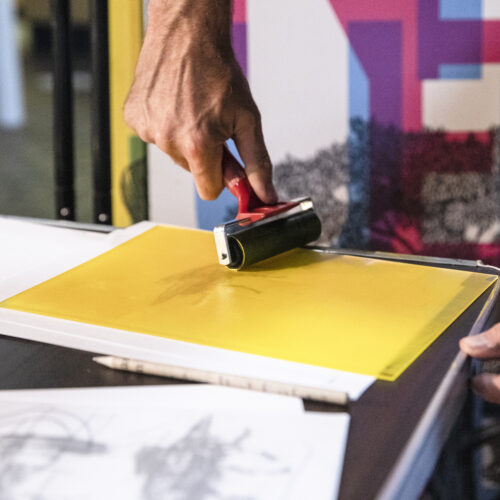 Hands rolling ink over a yellow perspex square placed over white paper