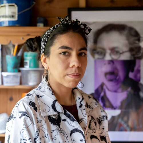 Mixed race woman with her hair tied back with a bandana wearing a white shirt with drawings of people's heads standing in front of a large collage portrait of a woman and some shelves full of pots