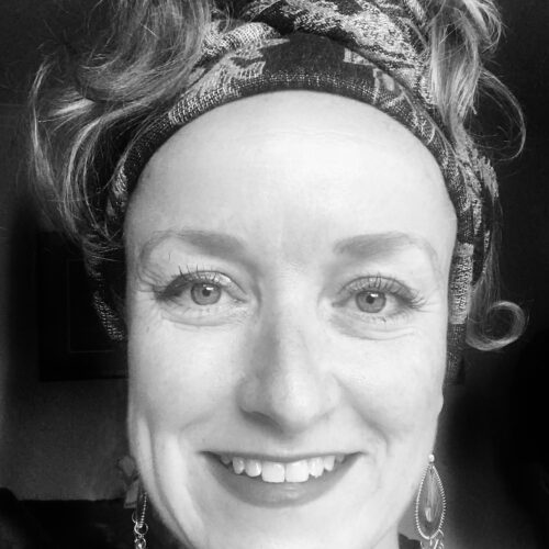 Black and white headshot of a white woman smiling at the camera, wearing her hair tied back by a patterned headband