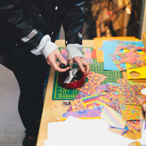 Hands making button badges using a small tool placed on a wooden work bench covered in patterned materials and pencils