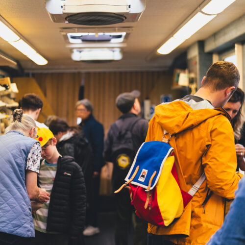 People stood in a bus decked out as a workshop. One man is wearing an orange coat and multi-coloured rucksack in blue, yellow and red. An older woman with grey hair is showing something to a young boy, who is wearing a yellow cap