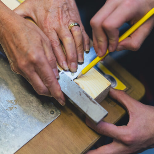 Two sets of hands, one younger than the other, holding a piece of wood and metal. One hand is drawing on the wooden cube with a yellow pencil