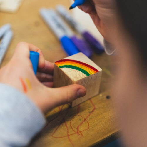 The hands of a child are drawing a colourful rainbow on a wooden block