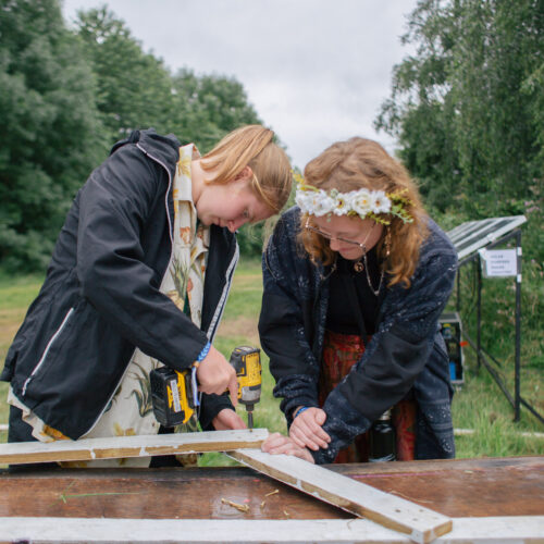 Two young red-headed girls are putting something together outside using wood. One girl wearing a daisy chain in her hair holds the wood as the other girl drills into the wood