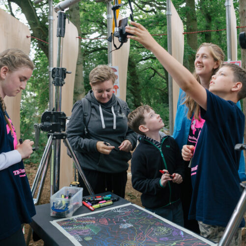 A group of young people and two adults looking at a large multi-coloured drawing on a table surround by camera equipment set up in a woodland